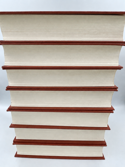 A stack of seven hardcover books with cream-colored pages and reddish-brown spines, photographed against a light gray background.