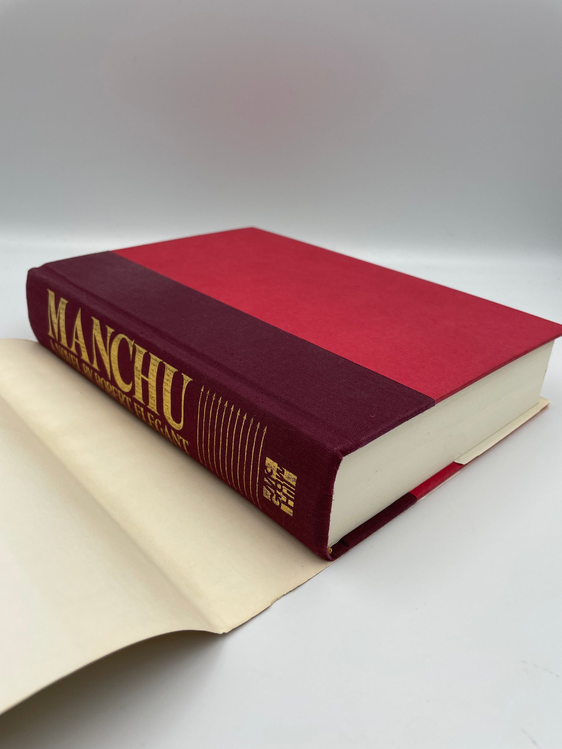 Book titled Manchu by Rober Elegant without dustjacket with red and beige cover on a white background.