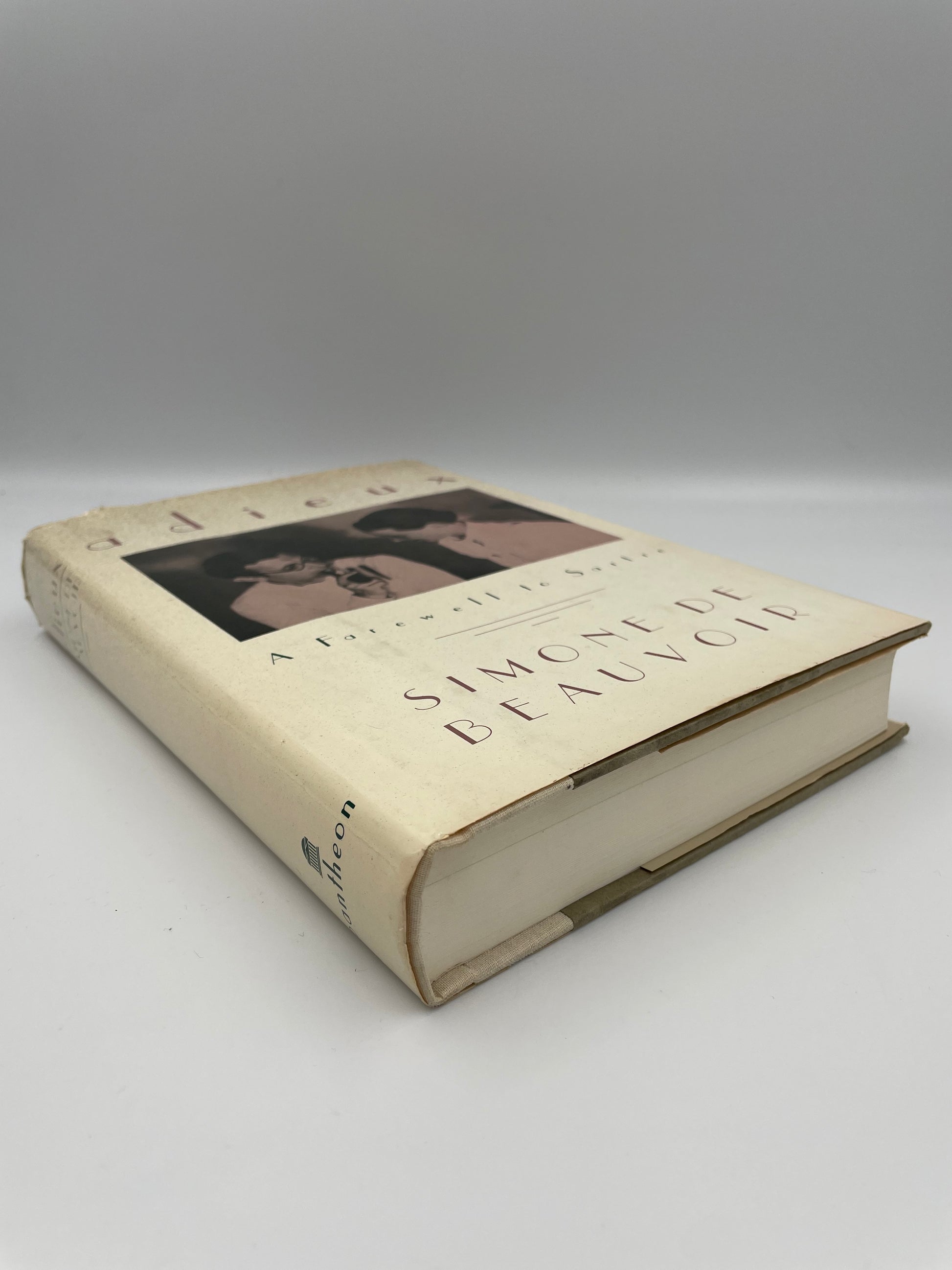 Book entitled ‘Adieux: A Farewell to Sarte’ by Simone de Beauvoir lying on its side with text and images on a white background. 
